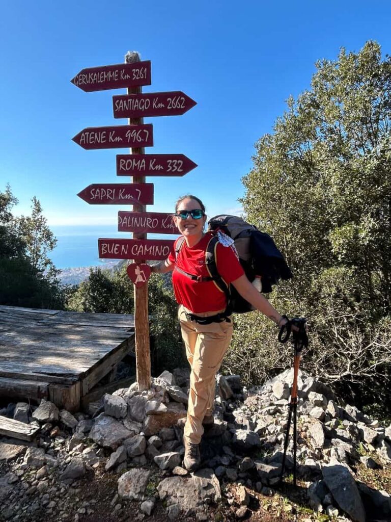 Camminatrice travel designer lungo il Cammino di San Nilo in Cilento, che posa in attrezzatura da trekking vicino a un palo della segnaletica con vista sul mare del Cilento